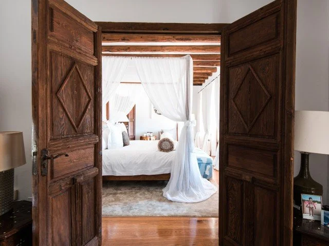 View through wooden double doors into a bedroom with a canopy bed with white drapes, wooden ceiling beams, and neutral decor.