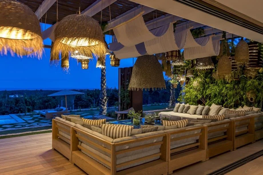 Indoor lounge area with wooden furniture and striped cushions, decorated with hanging woven lamps, and outdoor pool with umbrella visible through large open window at dusk.