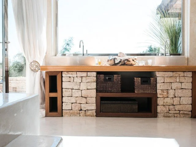 A kitchen with a stone brick counter, wooden surface, and open shelves, with a window showing outdoor plants in the background.