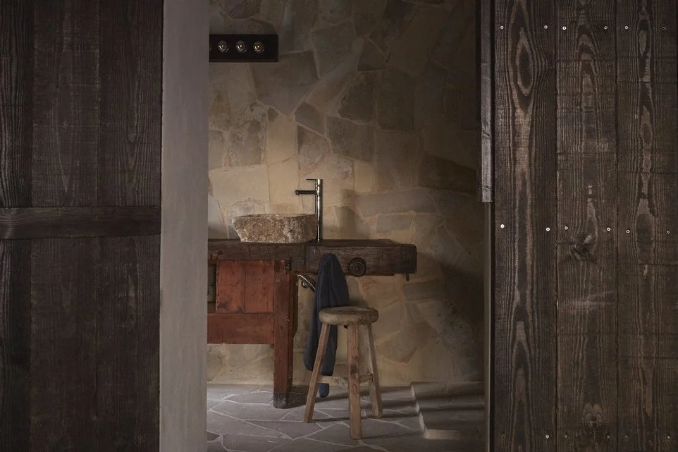 View of a rustic wooden bathroom with a stone sink, a wooden stool, and a dark towel hanging on the sink. There are stone walls and wooden sliding doors.