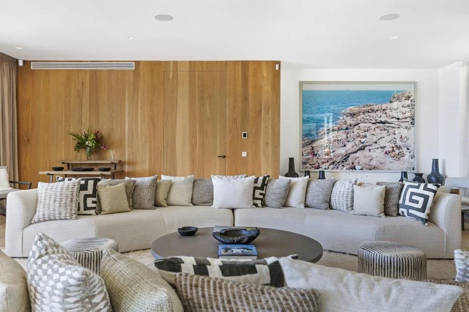 Modern living room with a large white sectional sofa adorned with various beige, gray, and patterned pillows. A round coffee table with black bowls and a book is in front. The wall behind has a wooden panel with a small table holding a flower arrange