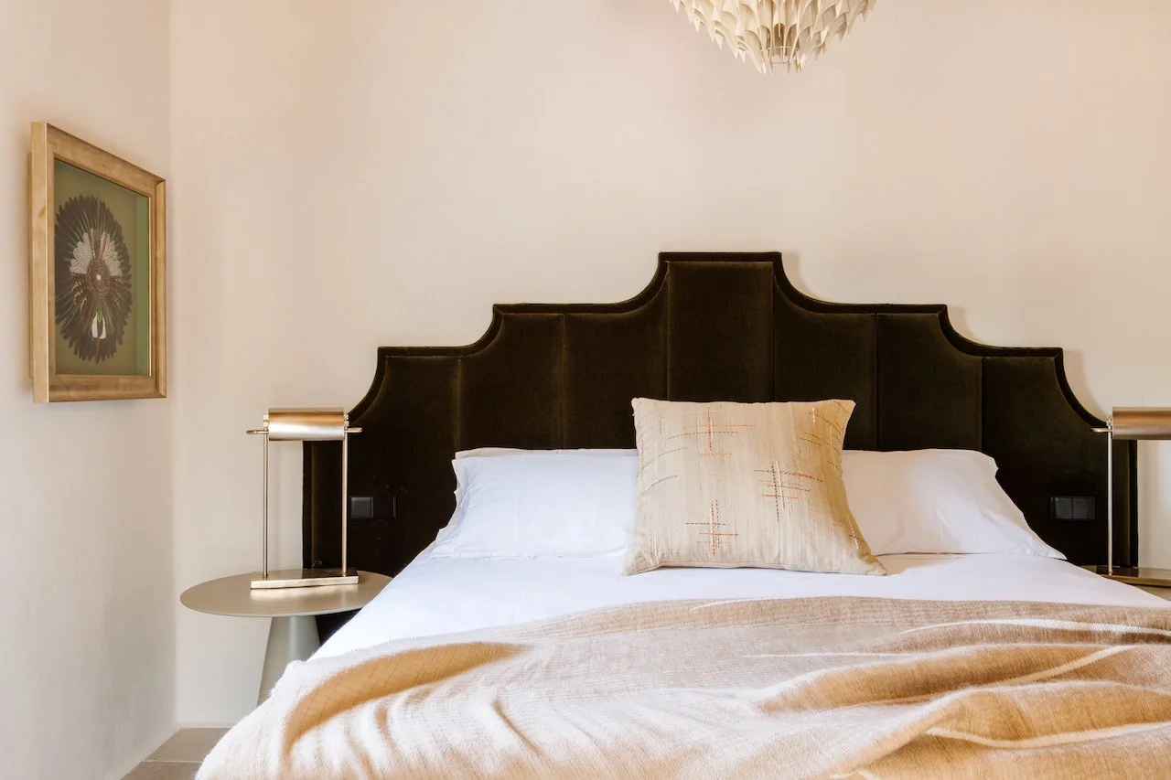 A neatly made bed with a black upholstered headboard, white linens, beige pillows, and a beige blanket in a bedroom with minimalist decor. There are two bedside tables with modern lamps and a framed artwork on the wall.