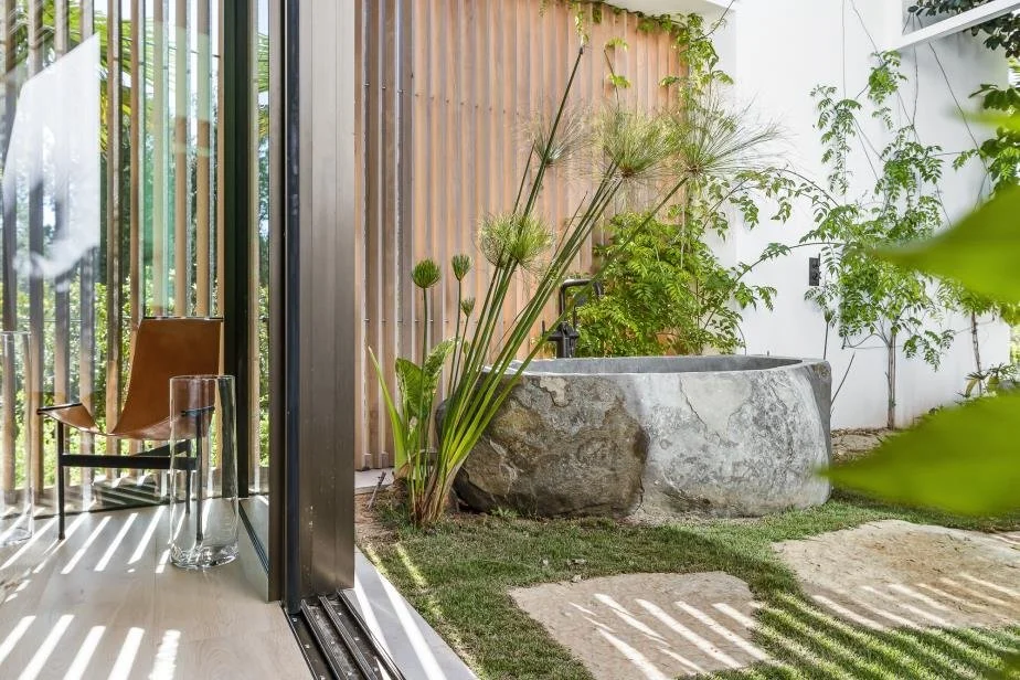 A modern indoor-outdoor space with a glass sliding door, a leather chair, glass table, and a large stone bathtub surrounded by green plants and natural light.