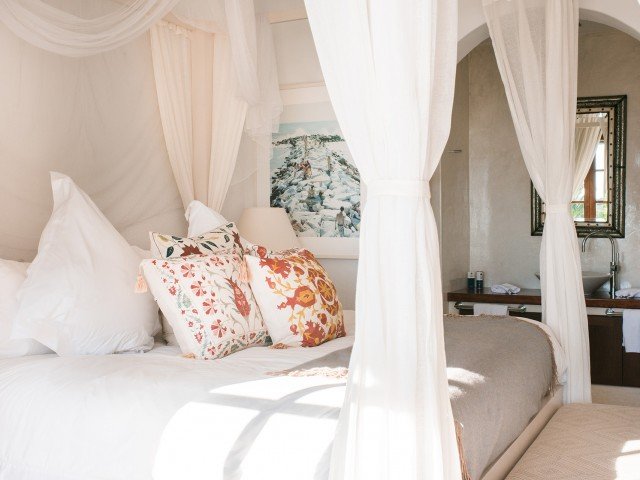 A cozy bedroom with white curtains surrounding a bed with white and patterned pillows, a beige bedspread, and a picture hanging on the wall behind the bed. A small vanity or sink area is visible in the background.
