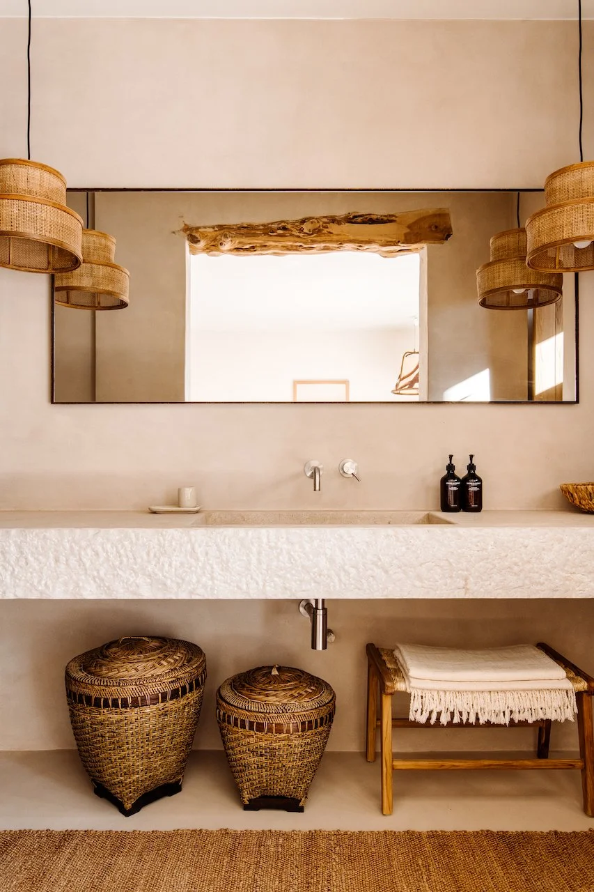 A bathroom vanity with a textured white countertop, a large rectangular mirror, and two wicker baskets below. There are two black soap dispensers on the right side of the counter and a small cup with a saucer on the left. Two woven pendant lights han