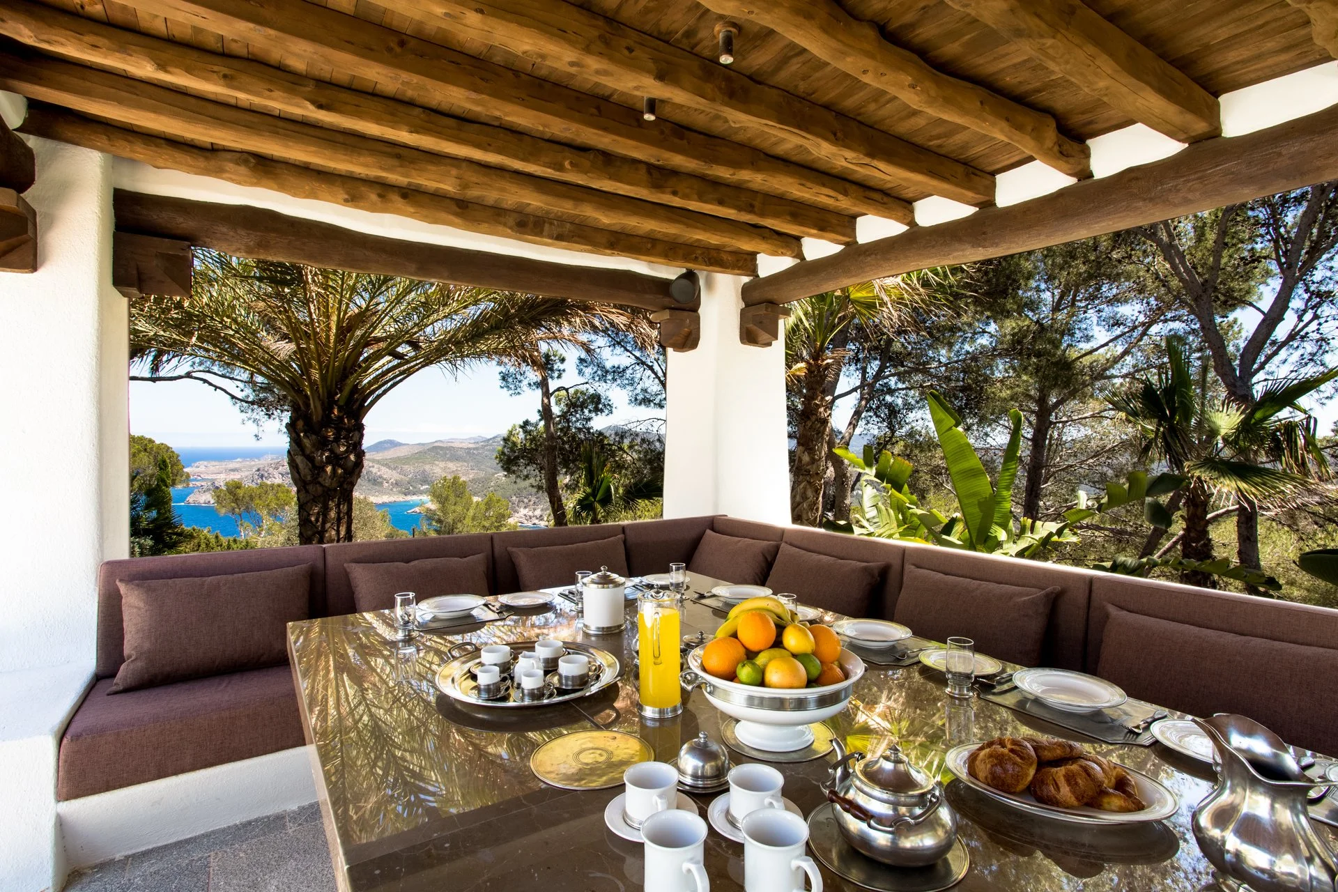 Outdoor dining area with a glass table set with coffee cups, a fruit bowl, croissants, and beverages. Surrounded by a brown cushioned bench, with a scenic view of trees, mountains, and ocean in the background.