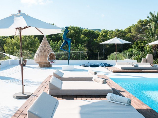 Luxury poolside area with lounge chairs, umbrellas, cushioned seating, and a modern sculpture with greenery in the background.