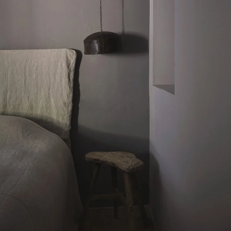 A bedroom corner with a bed, a small wooden stool, and a hanging light fixture.