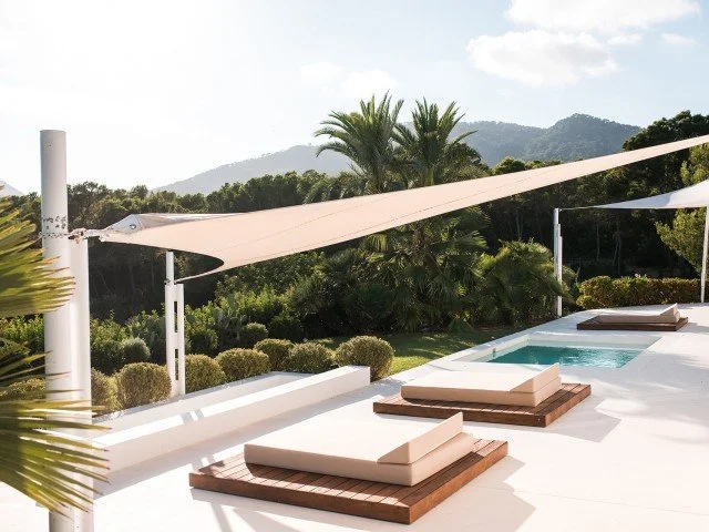 Outdoor poolside lounge chairs with cushions and wooden base, shaded by sail shades, overlooking a lush green landscape with trees and mountains in the background.