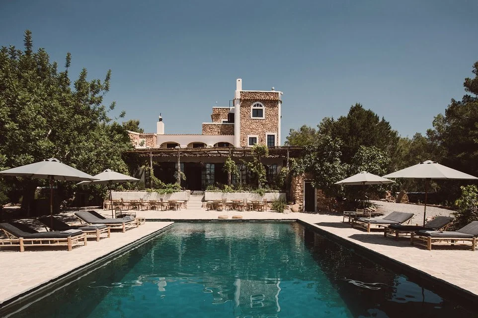 A luxurious outdoor swimming pool surrounded by lounge chairs with umbrellas, and a historic building with a tower and greenery in the background.