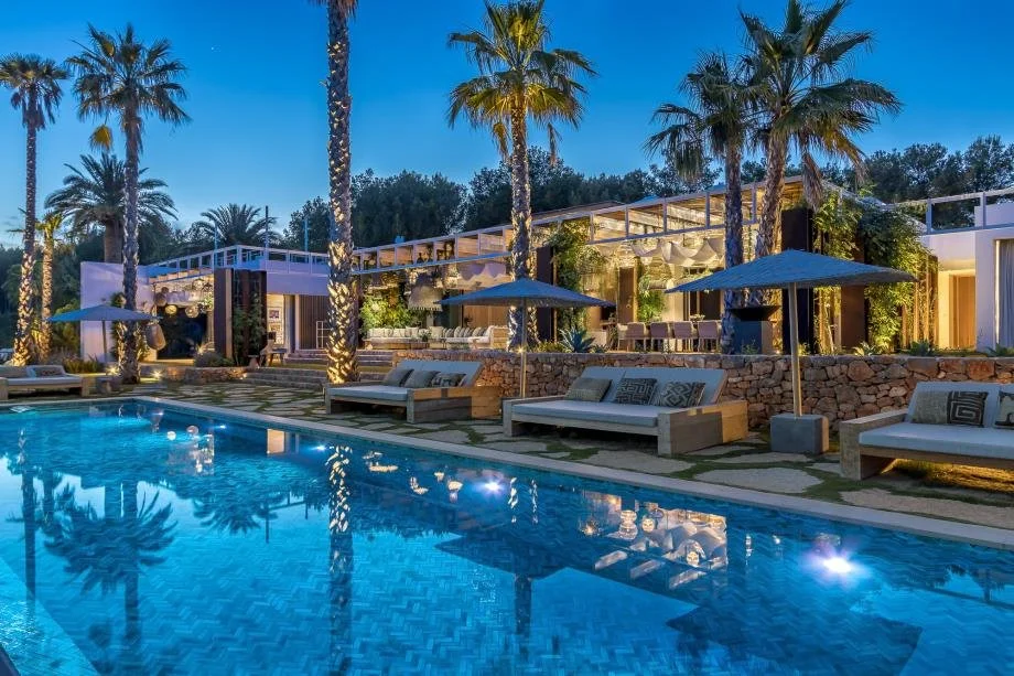 Outdoor pool area with lounge chairs, umbrellas, palm trees, and a modern building with lighting, set in the evening.