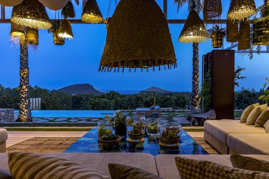 Indoor seating area with a table holding small glass terrariums, overlooking a pool and scenic landscape with trees, mountains, and a clear blue sky at dusk.