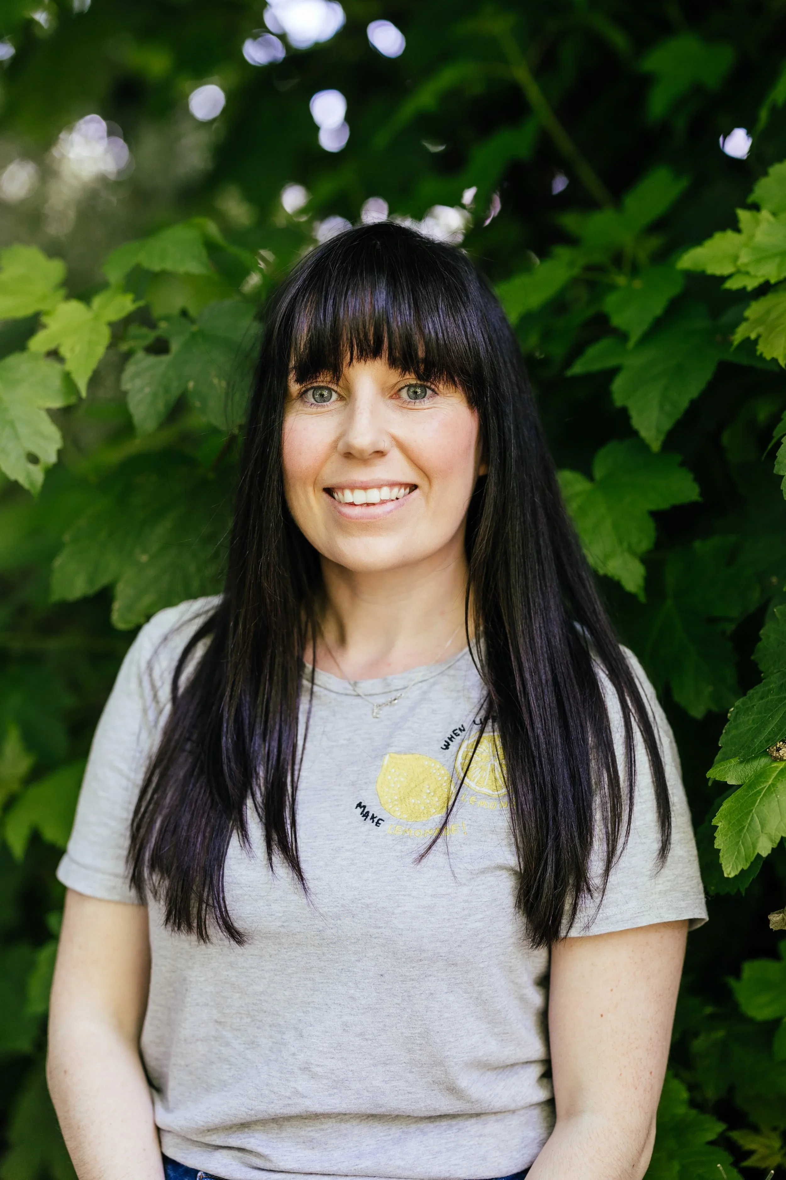 A woman with long black hair and blue eyes smiling outdoors in front of green leafy bushes.