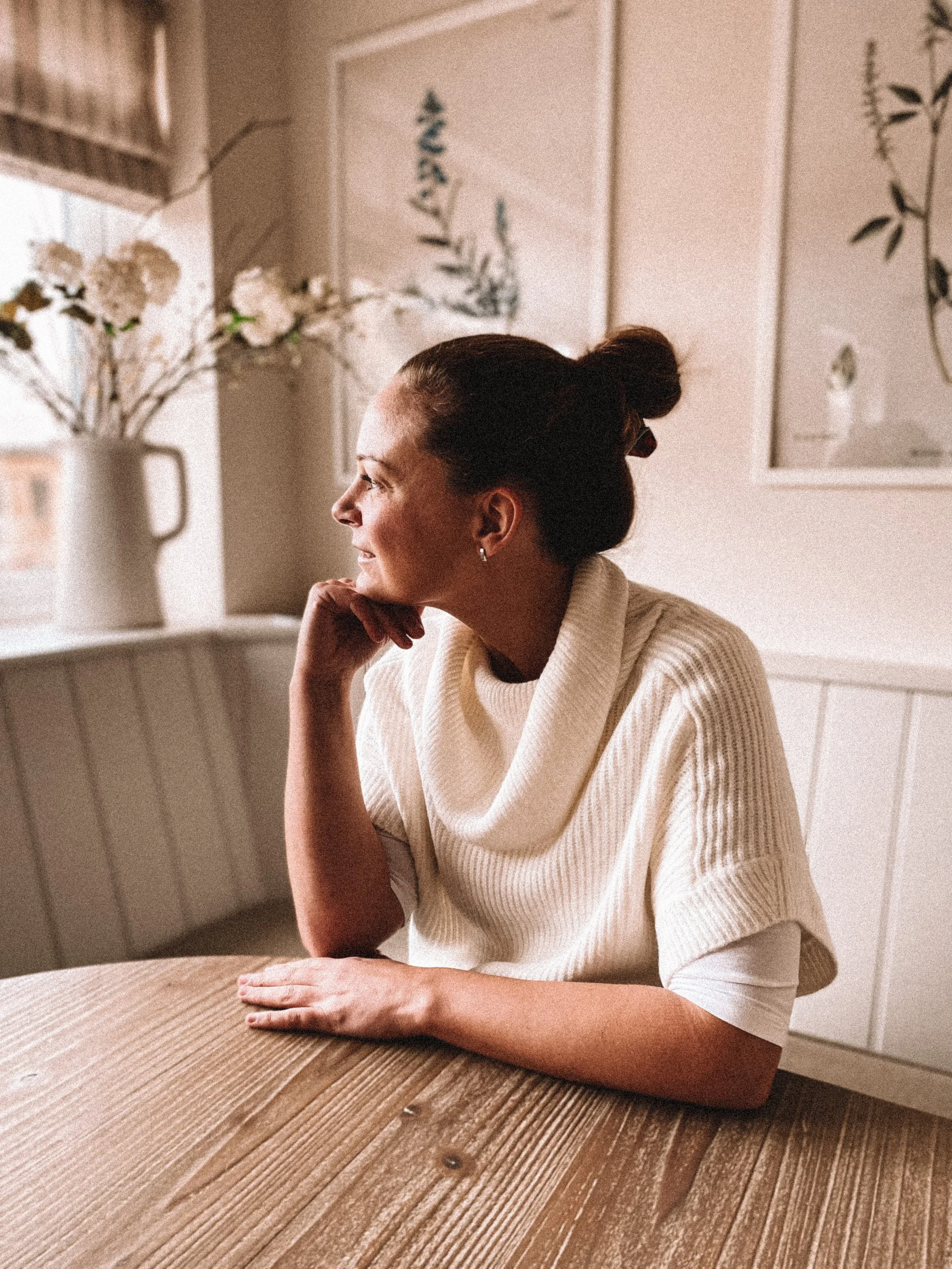 Woman sitting at a wooden table, looking out a window with a serene expression, wearing a cream-colored sweater.