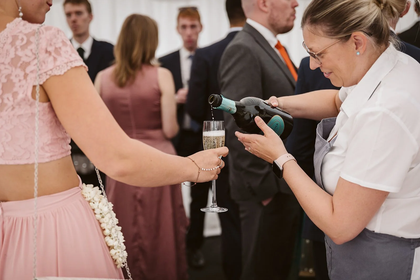 A woman in a white shirt pours champagne into a glass held by another woman in a pink dress at a social gathering.