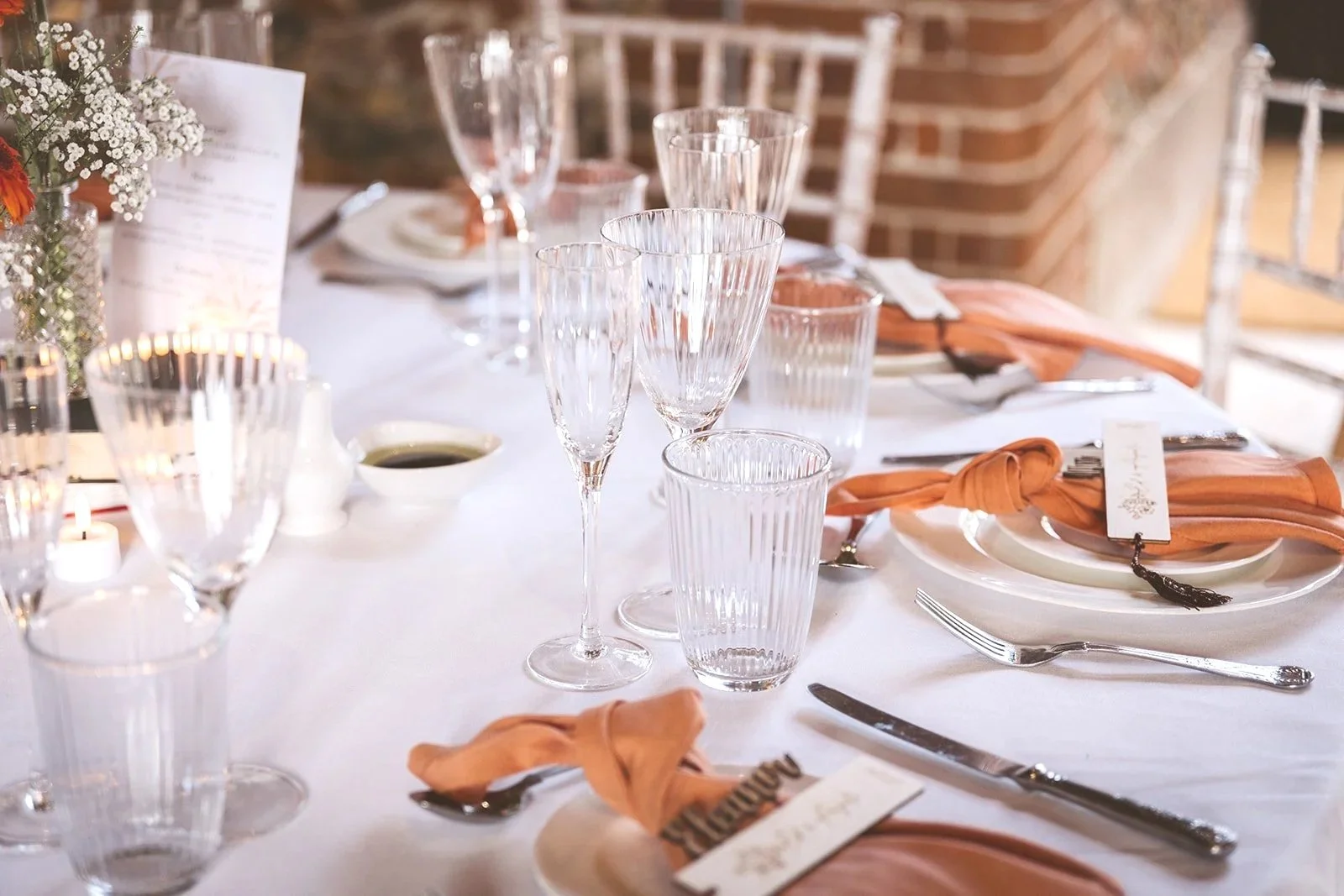 A formal dining table set with glassware, plates, silverware, and orange napkins, with a centerpiece of flowers and candles.