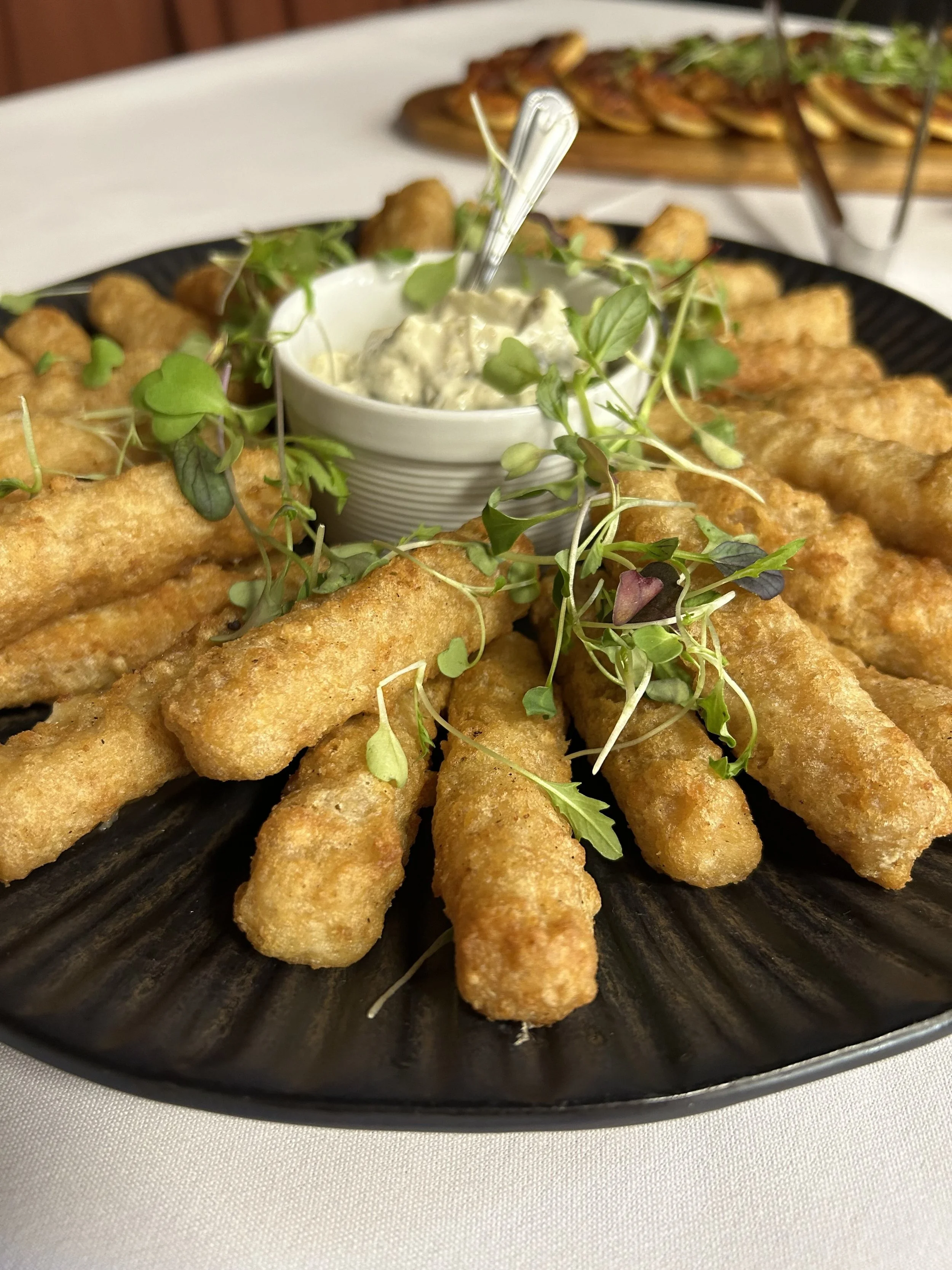 Fried fish sticks garnished with microgreens served with tartar sauce on a black plate.