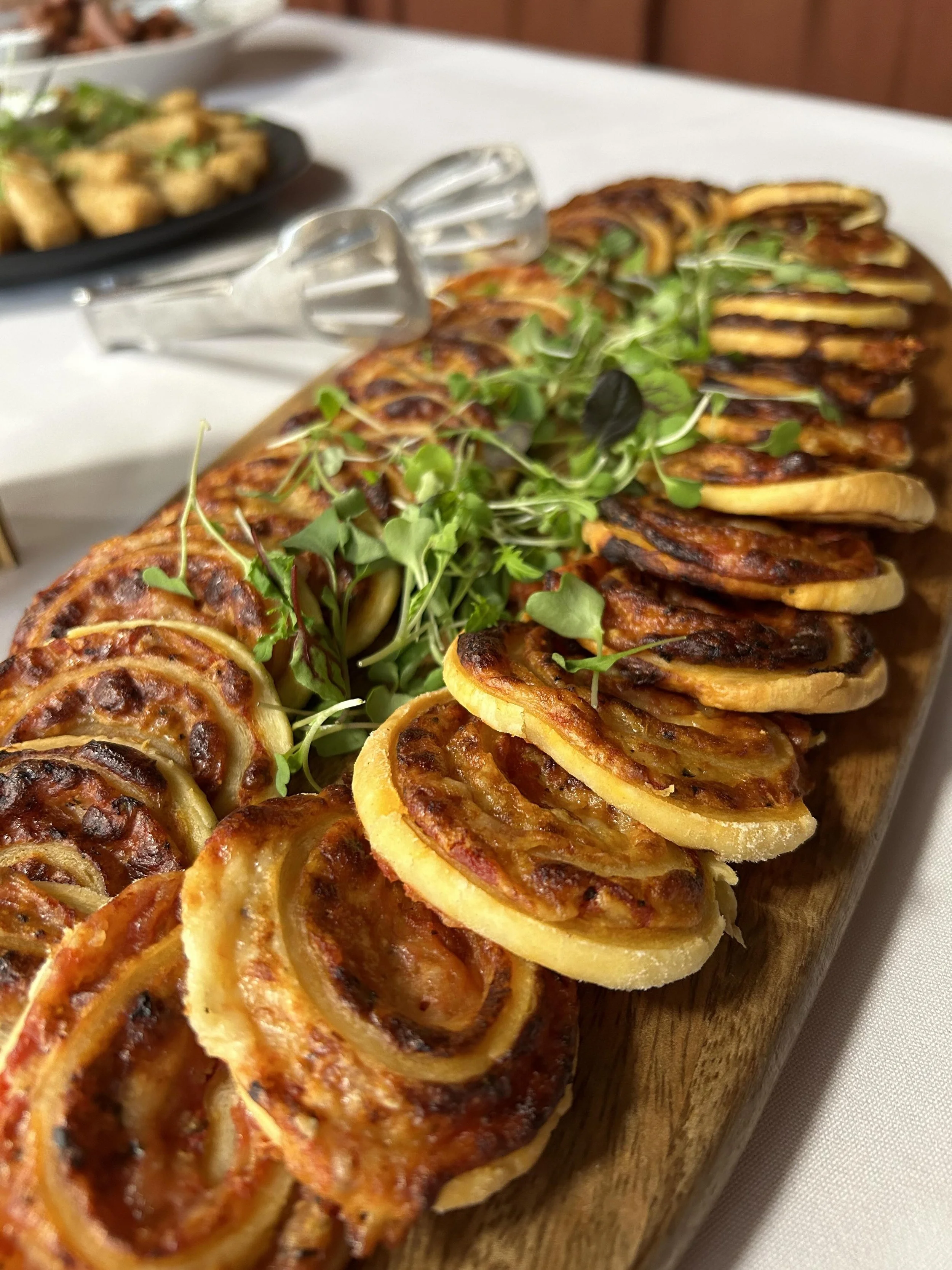 Close-up of a wooden platter with spiral-cut, baked stuffed vegetables, garnished with microgreens on a white table.