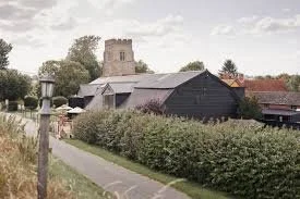 A pathways leading through greenery with bushes, an old black barn, and a church in the background under a cloudy sky.