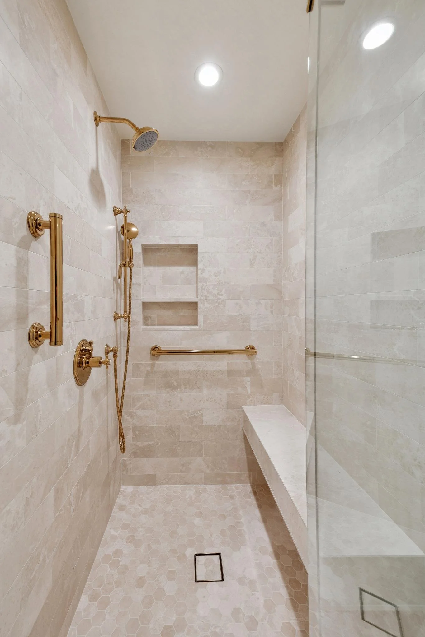 A walk-in shower with beige tiled walls and floor, gold fixtures including a showerhead, handheld shower, grab bars, built-in wall niches, and a bench.
