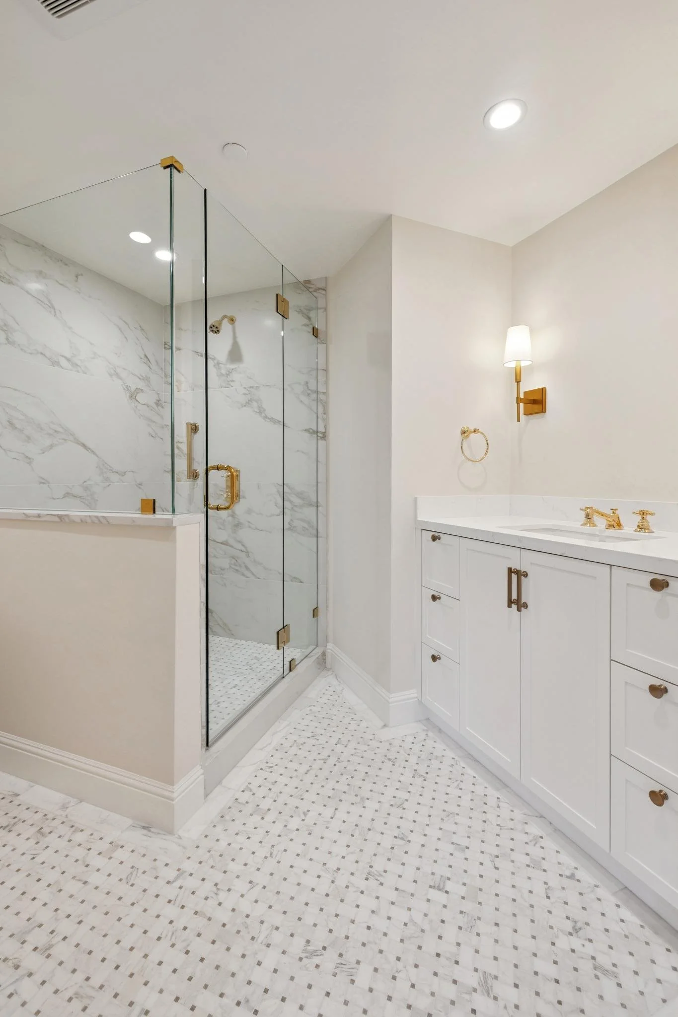 A modern bathroom featuring a glass-enclosed shower with white marble walls and gold fixtures, white cabinetry with gold hardware, a white countertop with a gold faucet, and a white tile floor with a small black diamond pattern.
