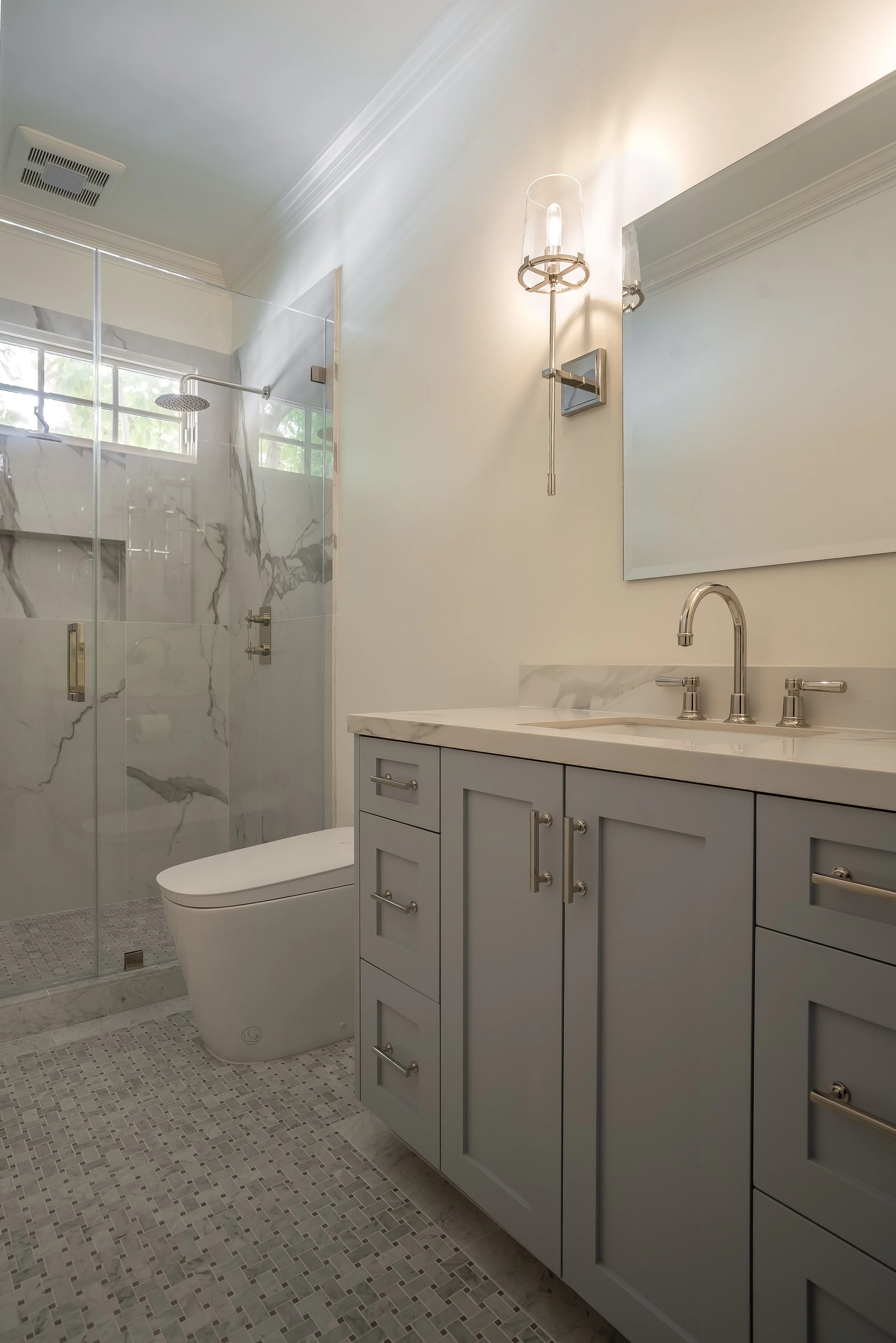 Modern bathroom with gray vanity, marble countertop, large mirror, wall-mounted light fixture, glass-enclosed shower with marble tile, and small mosaic tile flooring.