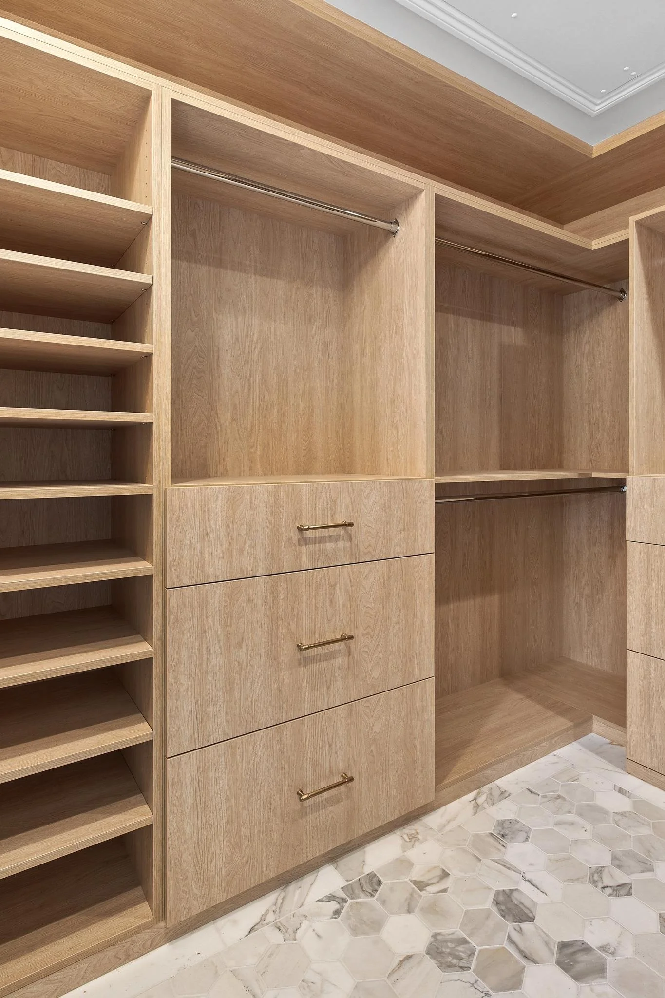 Empty wooden closet with shelves, drawers, and hanging rods, located on a hexagonal marble tile floor.