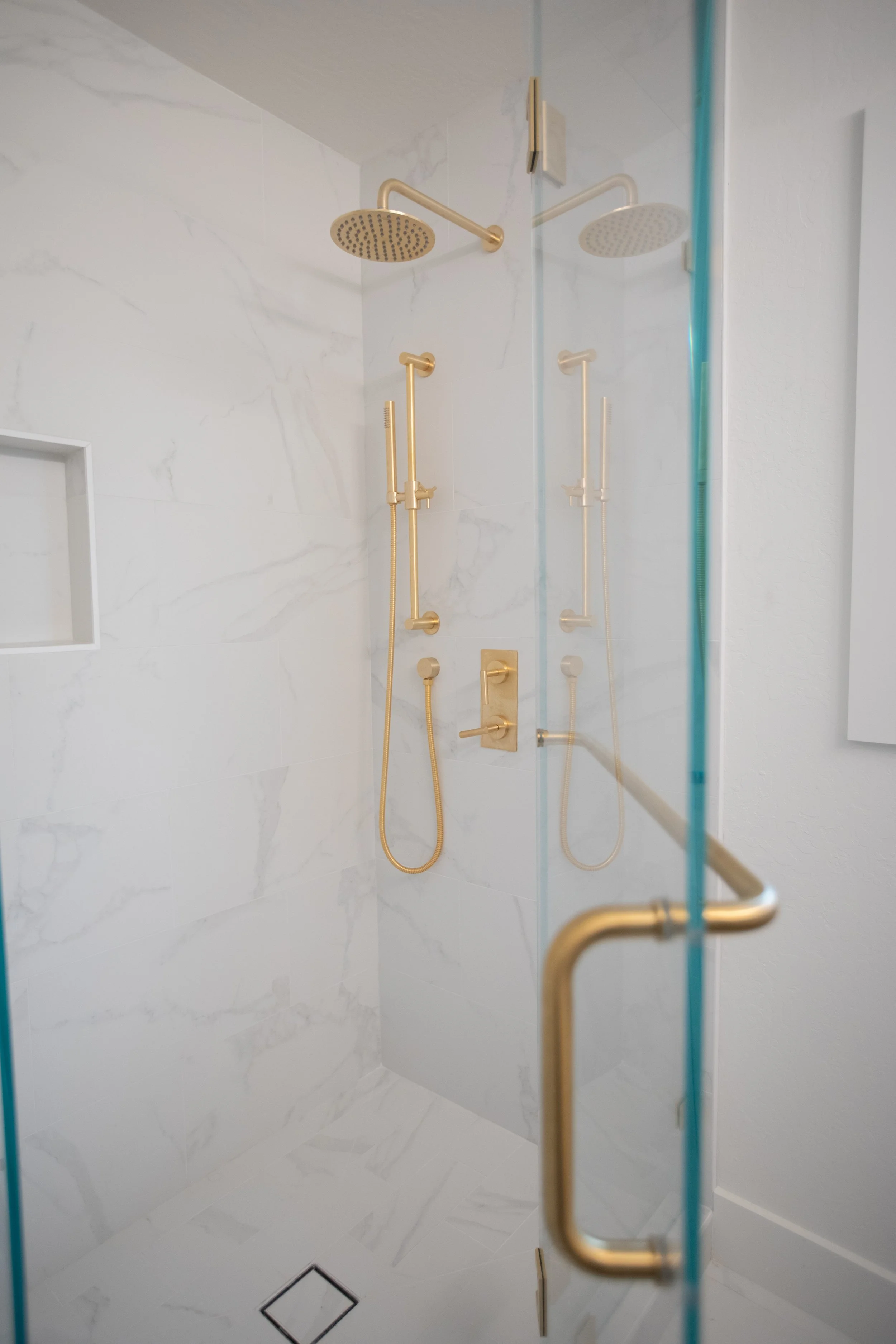 A modern walk-in shower with white marble tiles and gold fixtures, including two overhead rain showerheads, two adjustable handheld shower heads, and a built-in wall niche.