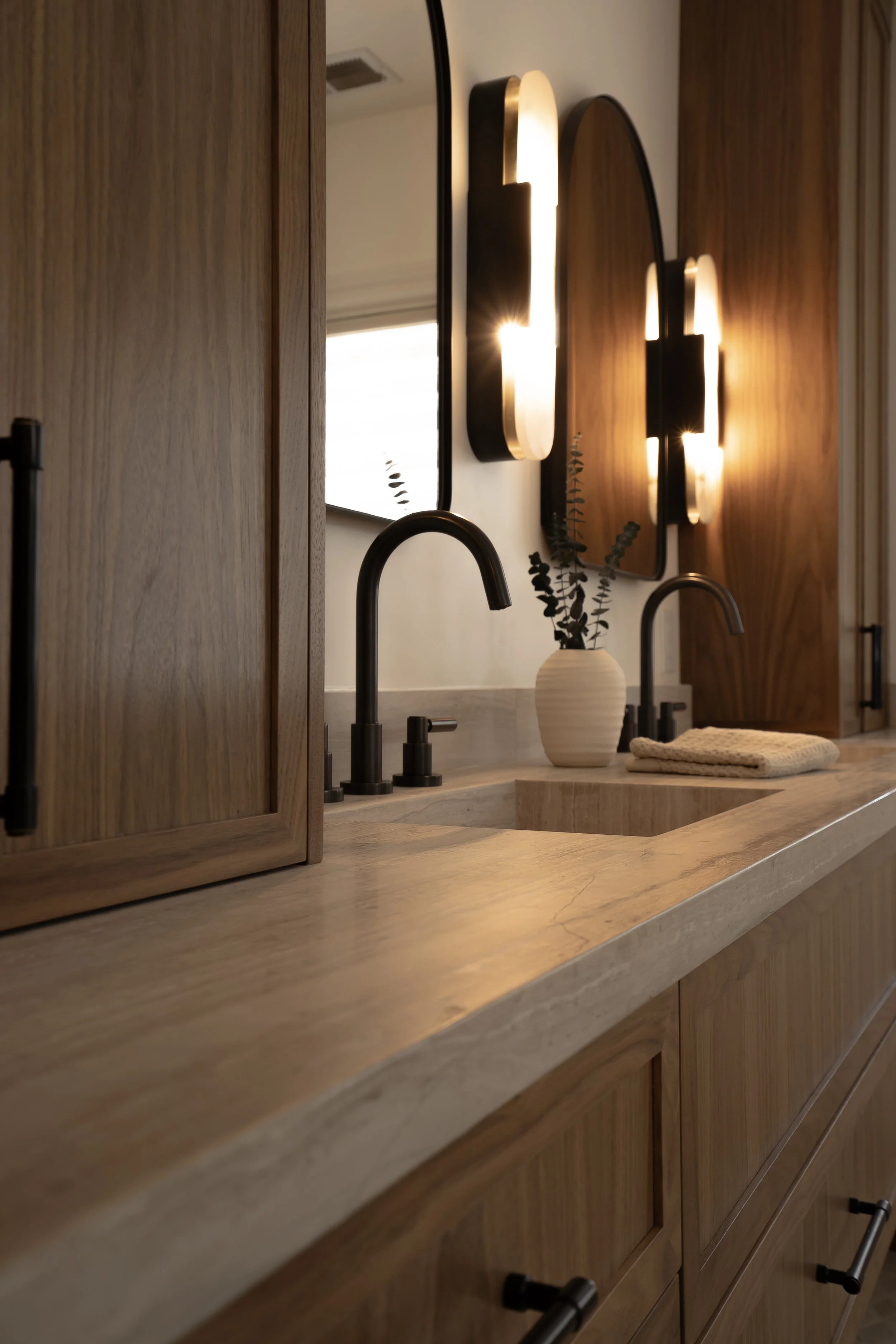 A modern bathroom vanity with a beige marble countertop, black faucets, a white vase with eucalyptus leaves, a towel, and two illuminated wall mirrors with black frames.