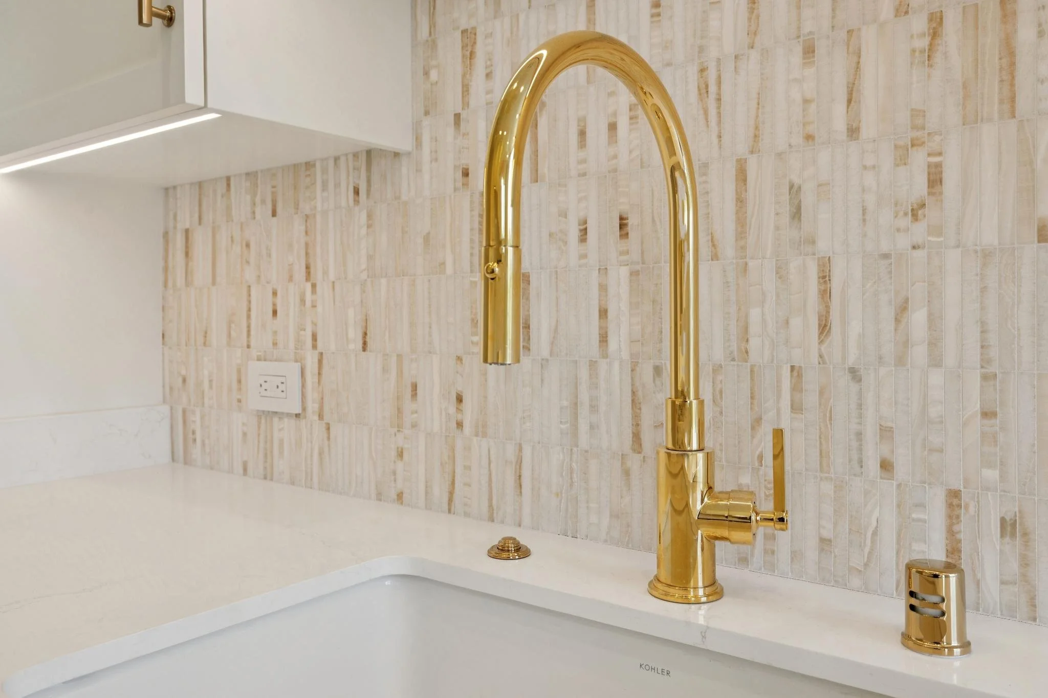 Close-up of a gold-colored kitchen faucet above a white sink with a marble countertop, beige tiled backsplash, and a wall electrical outlet.