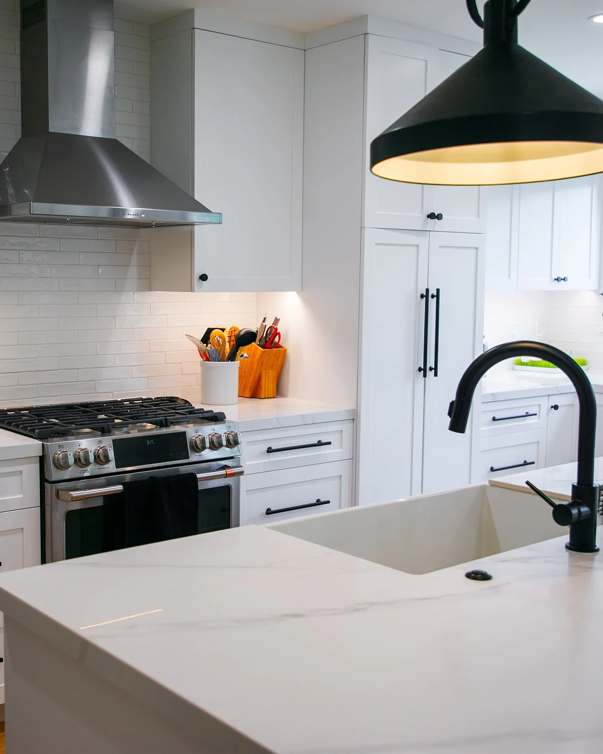Modern kitchen with white cabinets, black handles, stainless steel stove and range hood, black sink faucet, white marble countertop, and hanging black pendant light.