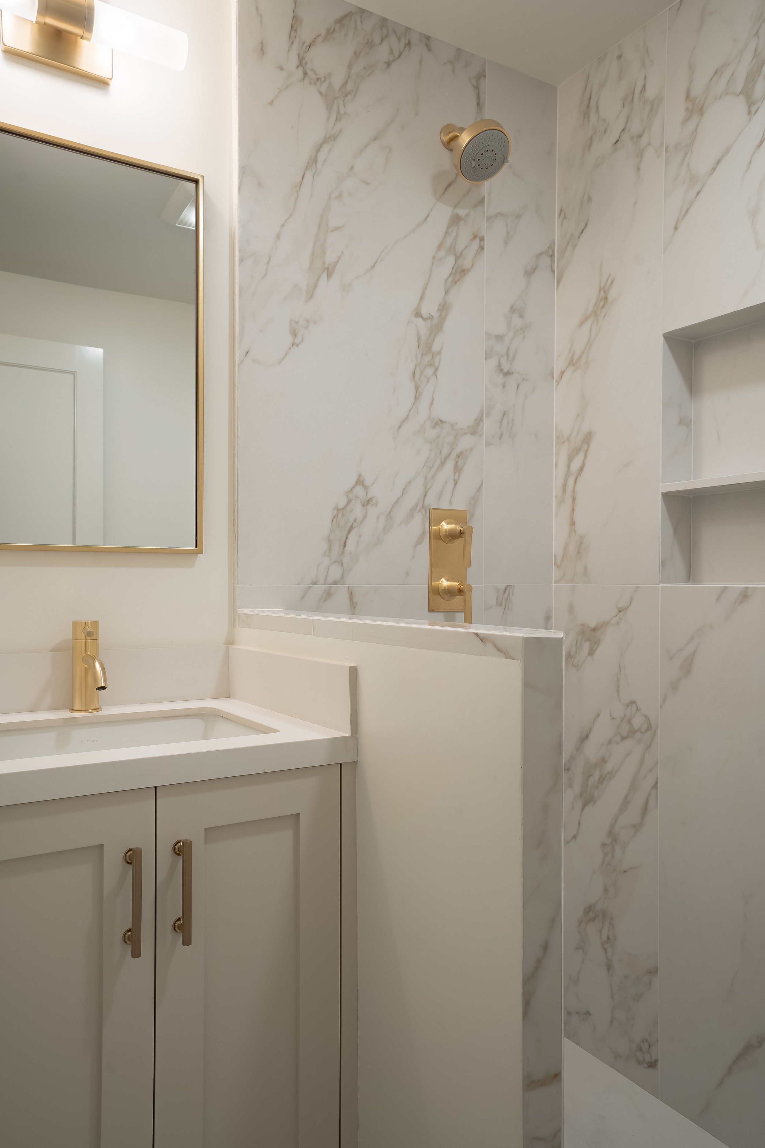 Modern bathroom with white and gold fixtures, a marble shower wall, a white vanity with a gold faucet, and a rectangular mirror.