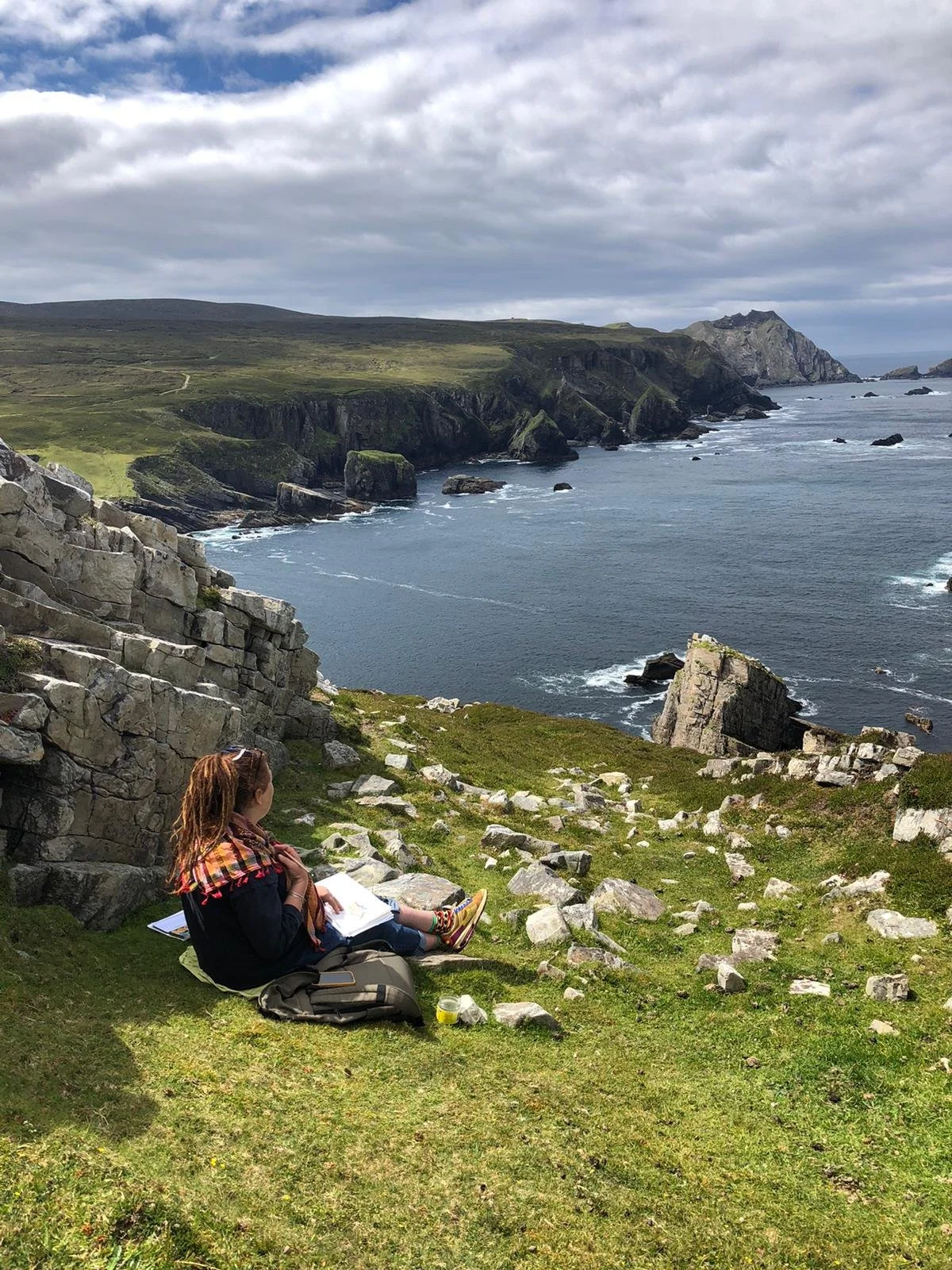 A person sitting on grass near rocky cliffs, overlooking the ocean with a cloudy sky overhead.