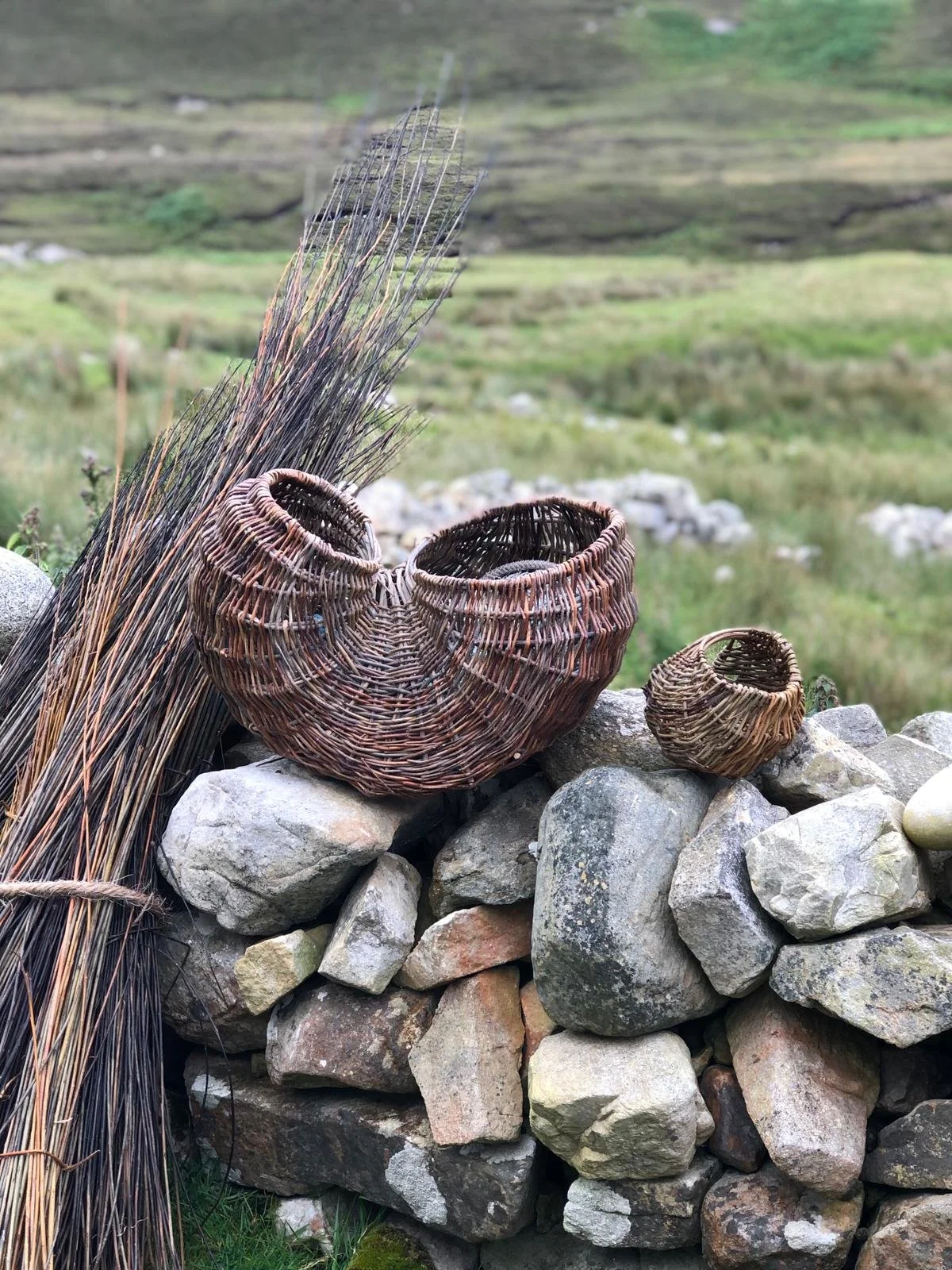 A collection of woven baskets and dried reeds resting on a stone wall in a rural outdoor landscape with green hills.