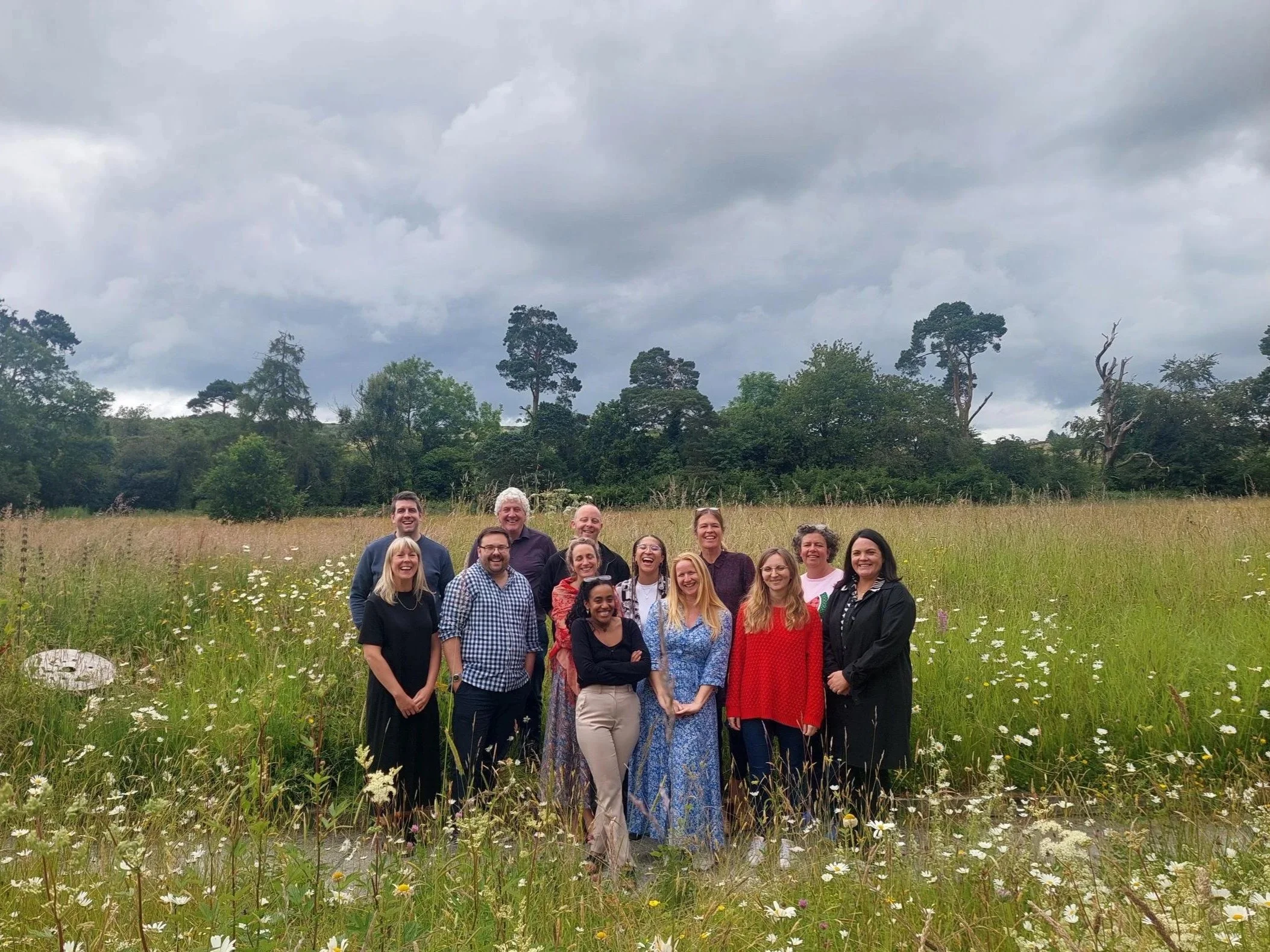 A group of people standing together outdoors in a grassy field with wildflowers, smiling. In the background are trees and a cloudy sky.