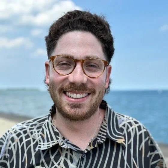 A smiling man with brown hair, glasses, and a beard, wearing a patterned shirt, standing on a beach with the ocean and blue sky in the background.