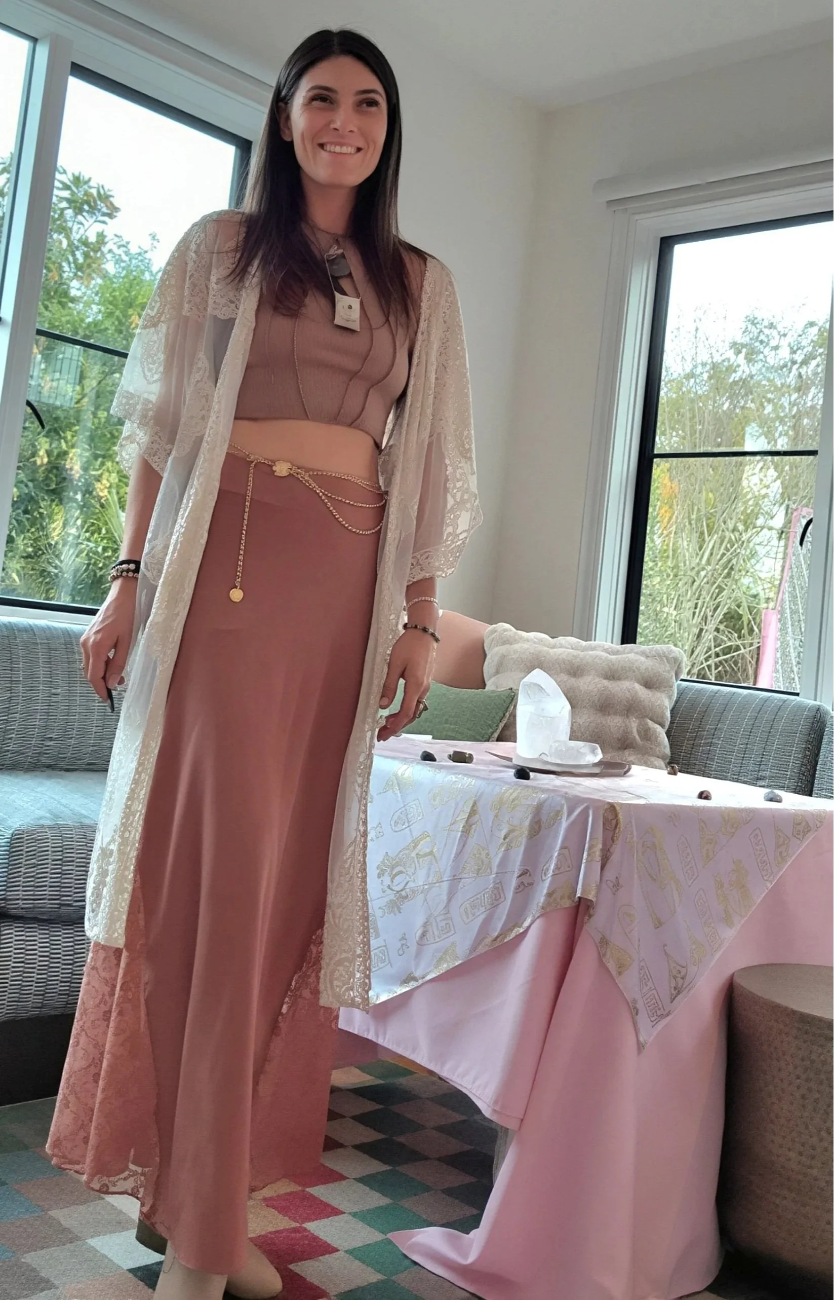 A woman with long dark hair wearing a peach-colored dress and lace shawl standing indoors near a window with greenery outside, smiling. There is a table with a pink tablecloth, decorative crystals, and small stones or jewelry, inside a room with a checkered carpet.