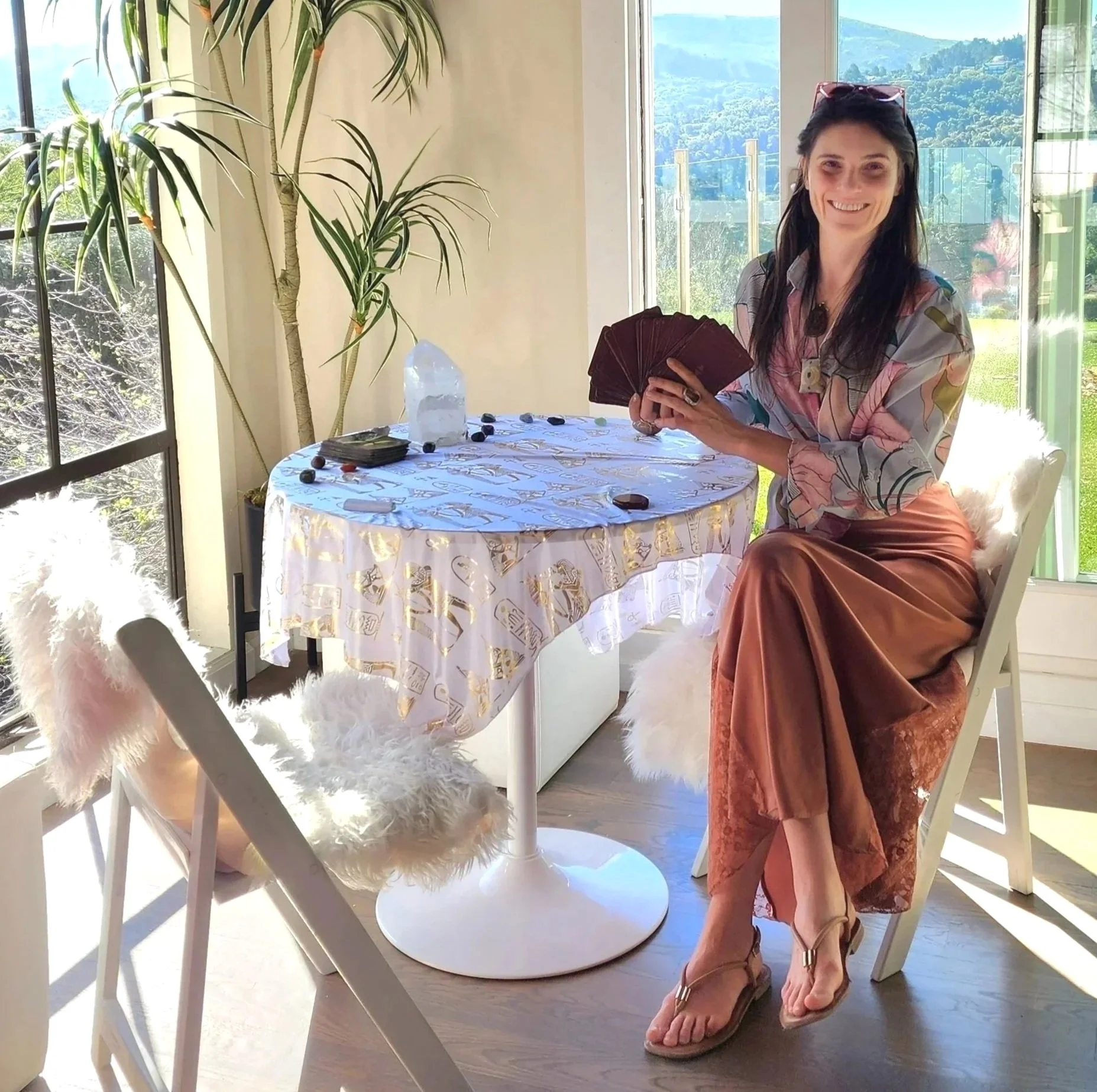 A woman smiling and holding a deck of tarot cards sits at a round table near large windows showing a scenic view of hills and trees. She is dressed in a colorful patterned top and a satin skirt, wearing flip-flops, with sunglasses on her head.
