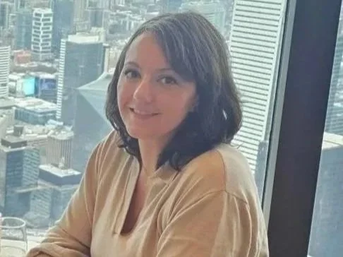 Woman with shoulder-length brown hair, wearing a beige top, sitting near a large window overlooking a city skyline.