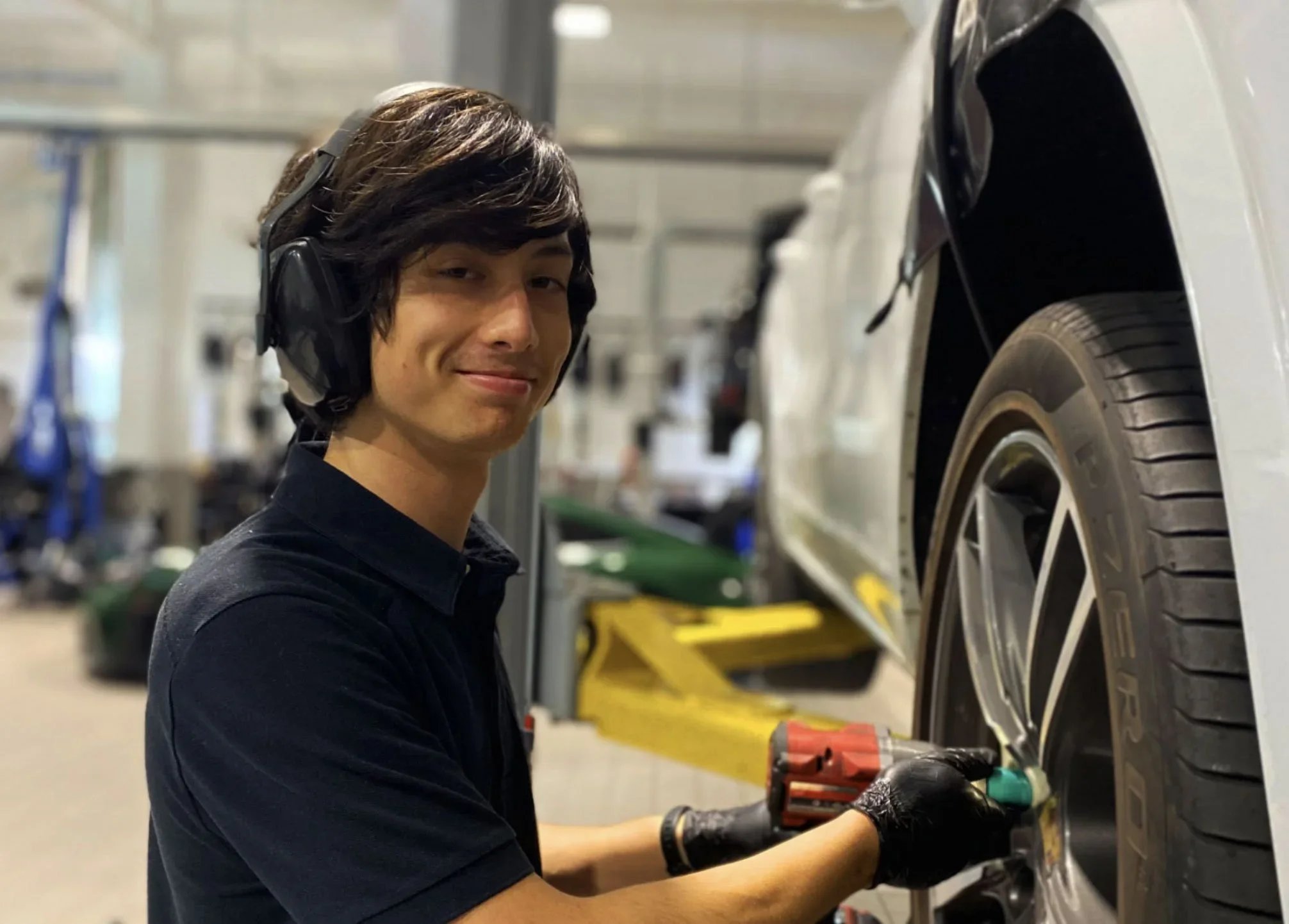 A mechanic wearing headphones and gloves is using a power tool to work on a car's wheel in an auto repair shop.