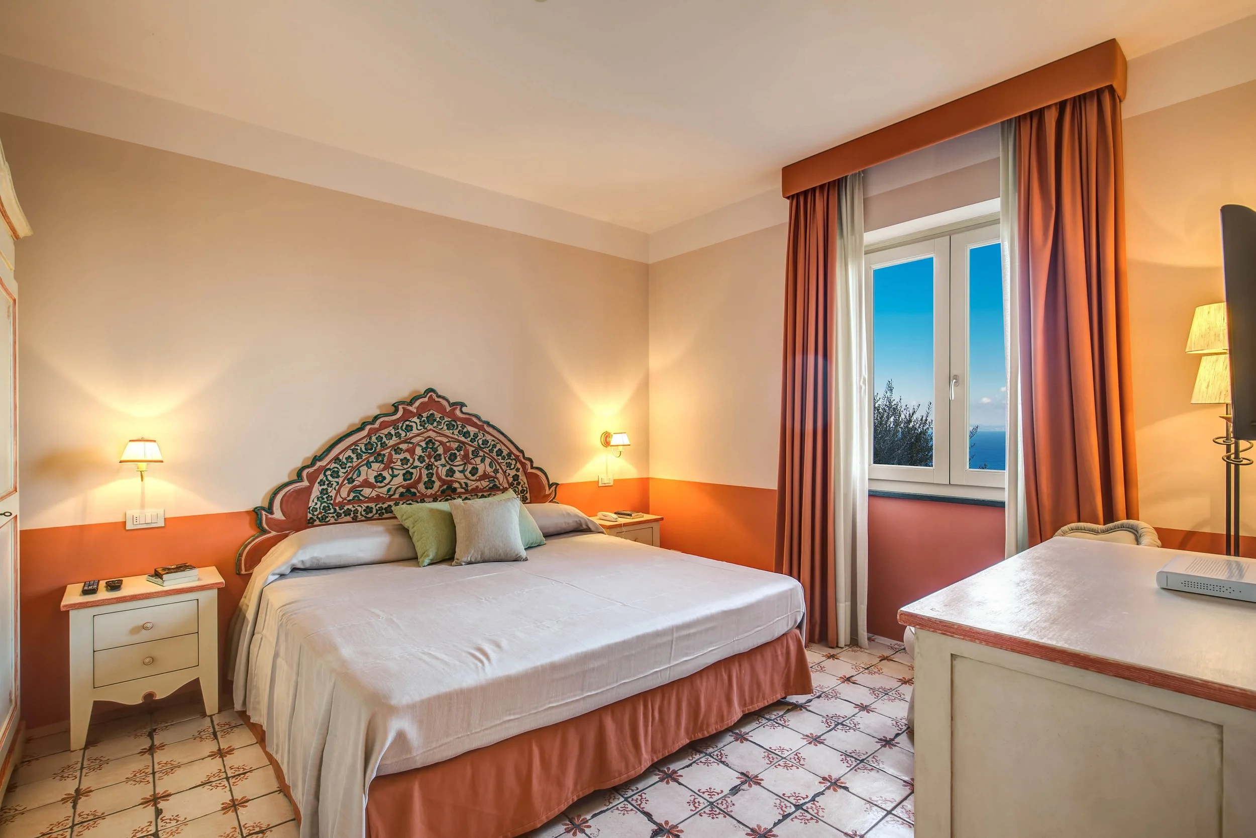 King size bed with detailed headboard in orange and white room with dresser, arm chair, and bright window with sea views in Villa Stradivari