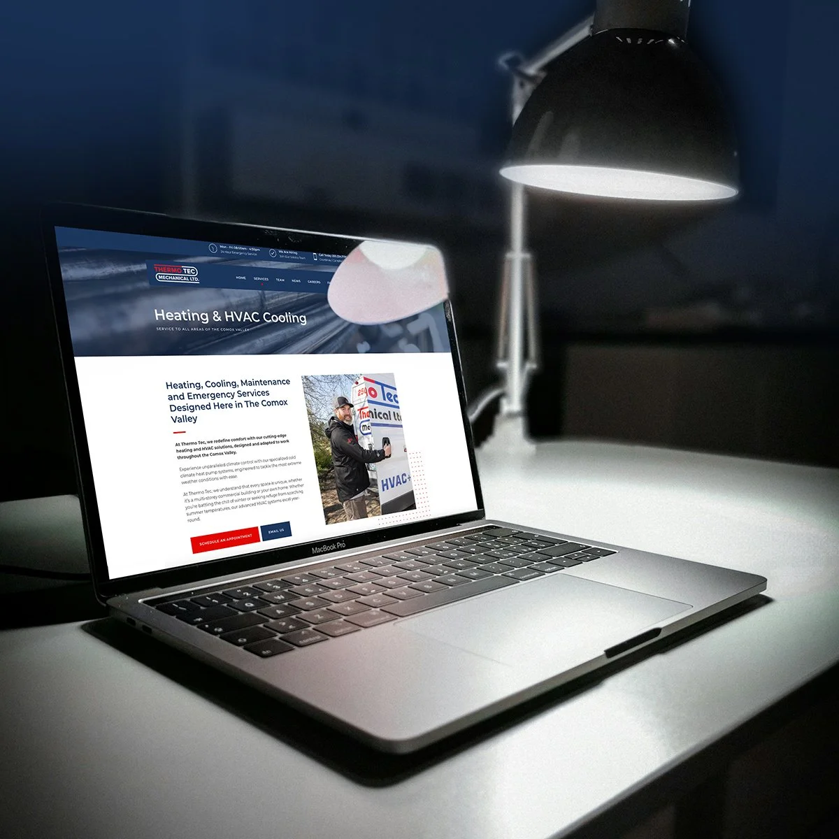 A laptop on a desk displaying a website for heating and HVAC cooling services, with a black desk lamp overhead in a dimly lit room.