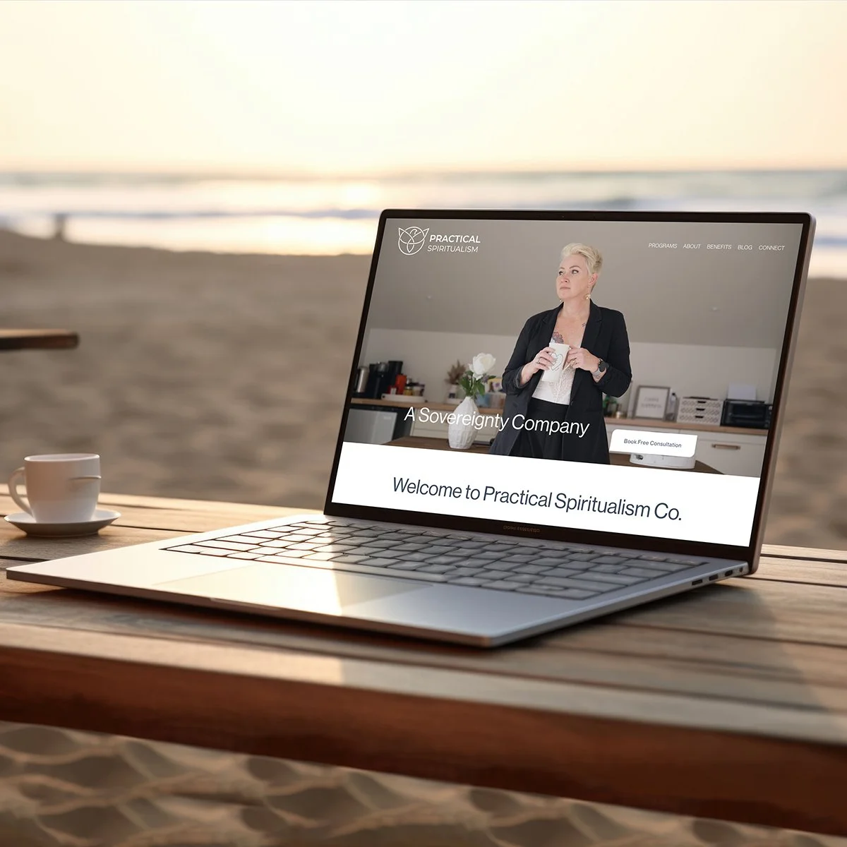 Laptop on a wooden table at the beach displaying the website of Practical Spiritualism Co., featuring a woman holding a cup in an indoor setting.