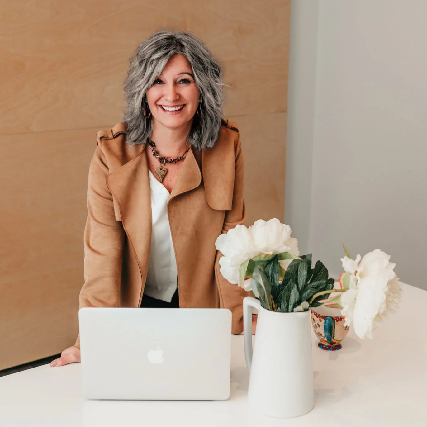 A smiling woman with gray hair stands behind a laptop on a table. She wears a tan coat over a white blouse. On the table is a white pitcher with white flowers and a colorful mug.