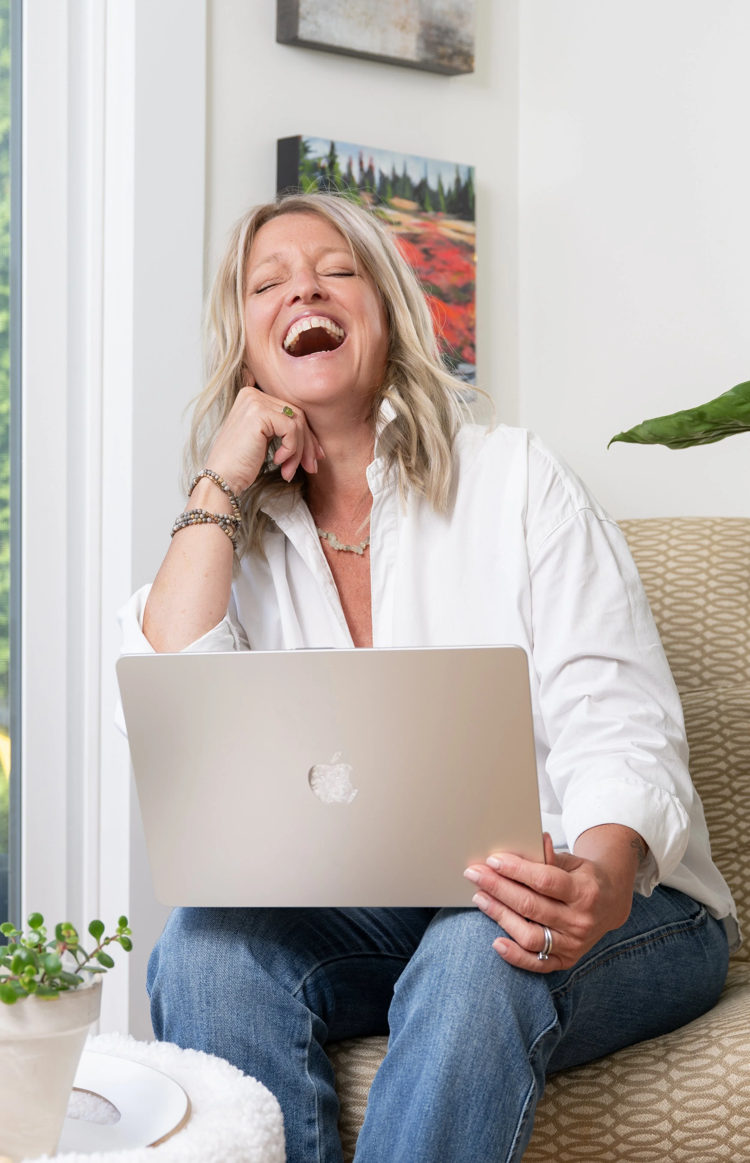 Chrissie laughing while sitting on a beige patterned chair, holding a silver MacBook laptop on her lap, with a window and artwork in the background.