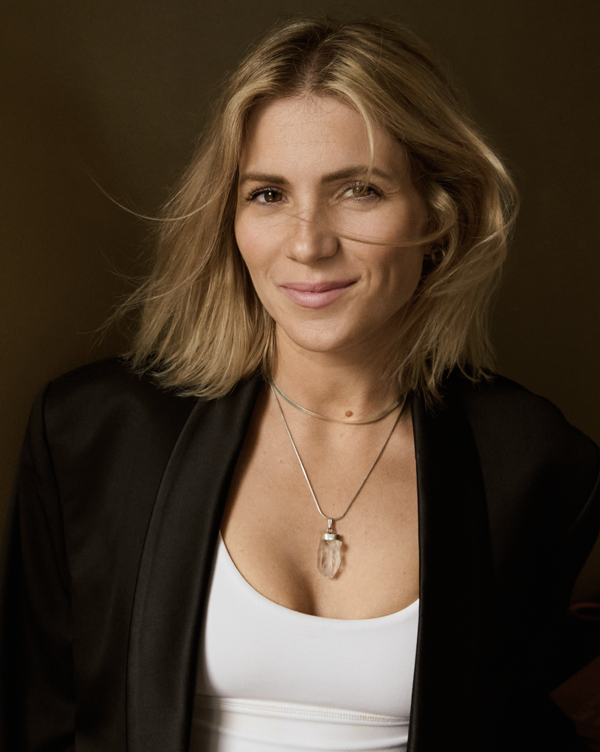Portrait of a smiling woman with blonde hair wearing a black blazer, white top, and a crystal pendant necklace, against a dark background.