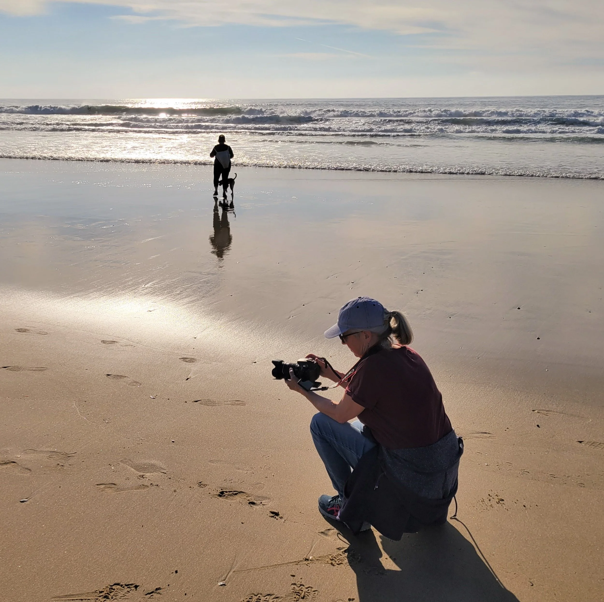 a photographer crouches to take a picture of a person walking a dog on the beach
