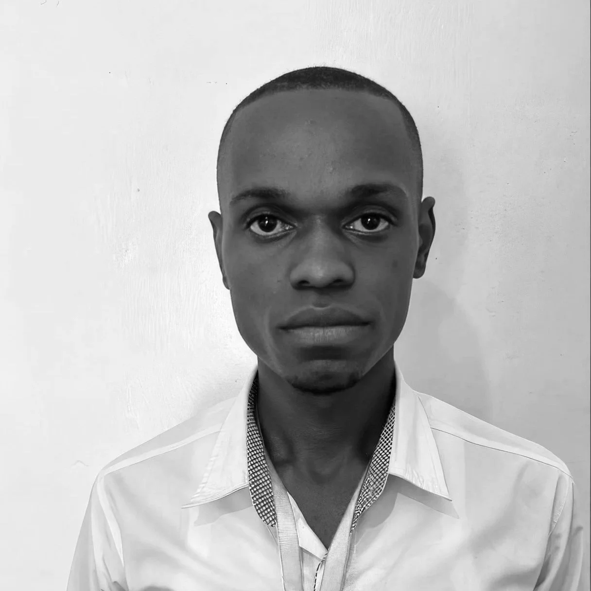 Black and white portrait of a young man with a buzz cut, wearing a collared shirt with a checked inner collar, standing against a plain light-colored wall.
