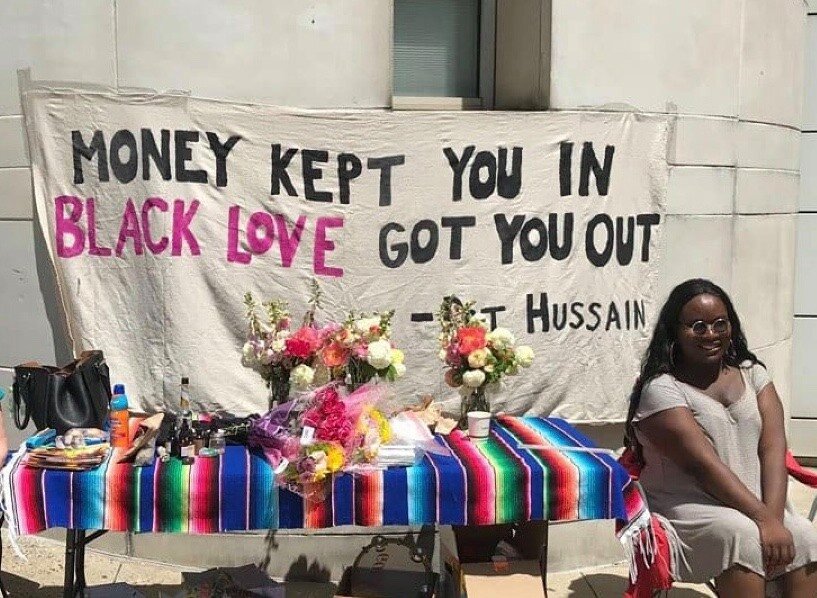 A photo of a person with sunglasses smiling and sitting on a bench in front of a sign saying “Money Kept You In Black Love Got You Out”.