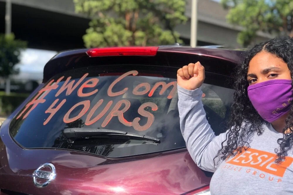 A photo of a person with their first up, curly hair, wearing an “Essie Justice Group” grey sweater and purple mask standing in front of a maroon car with “#We Come 4 Ours” painted on the back mirror.